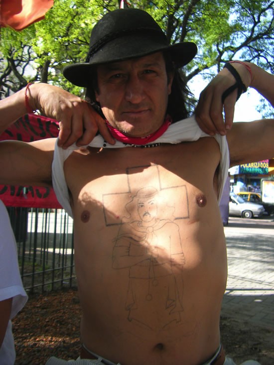 A man lifting up his shirt shows a crude Gauchito Gil tattoo on his chest. 
