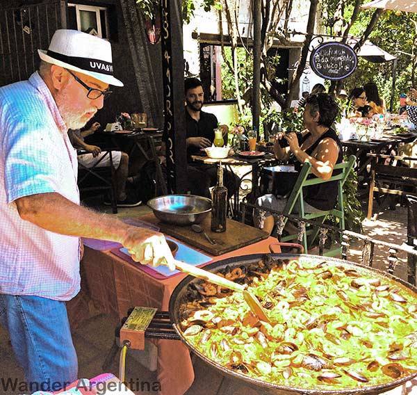 A man cooking a big pan of paella outdoors at a restaurant in Colonia del Sacramento, Uruguay,as customers look on from nearby tables. 