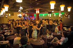 As people sit at tables in the foreground, a roomful of couples dance tango in the background at a Buenos Aires milonga, or tango dancehall
